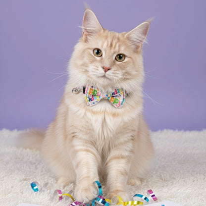 A fluffy cream-colored cat sitting against a soft purple background, wearing the Bling Fuzzie white cat collar with a colorful 'Happy Birthday' letter print and matching bow tie.