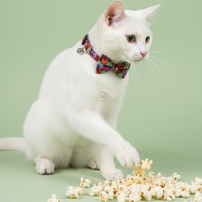 White cat wearing Bling Fuzzie Colorful Hearts Cat Collar with colorful bow tie and silver bell, playfully pawing at scattered popcorn on a sage green background