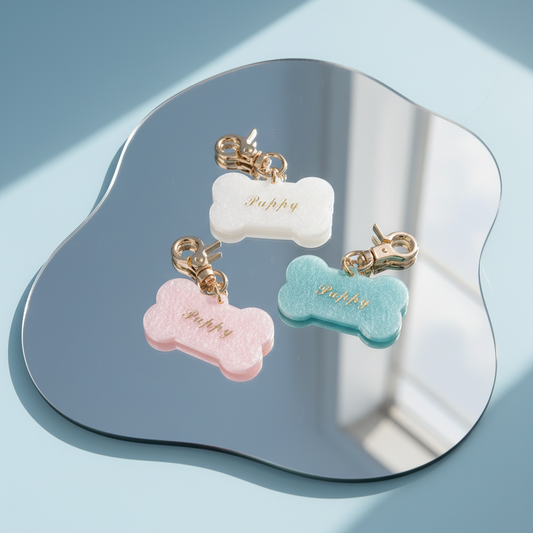 Three Bling Fuzzie Custom Dog Bone Pet ID Tags with gold foil engraving in white, pink, and teal glitter acetate, displayed on a mirror tray against a light blue background with natural shadow detail