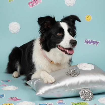 Black and white Border Collie resting on a silver satin cushion wearing a Bling Fuzzie blush pink tweed collar with gold brass hardware, surrounded by camellia flowers, glitter brooches, and colorful stickers against a light blue background
